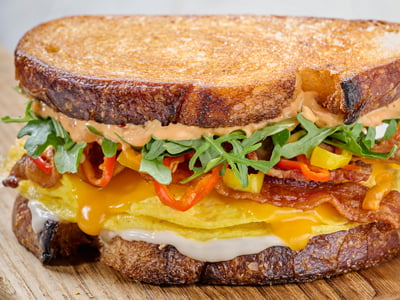 Bacon. Egg. Cheddar. Our twist on a traditional breakfast sandwich — hardwood smoked bacon, folded cage-free eggs, aged Cheddar, house-pickled sweet peppers and arugula with Calabrian chili aioli and roasted garlic aioli on griddled artisan sourdough bread. Served with lemon-dressed organic mixed greens.