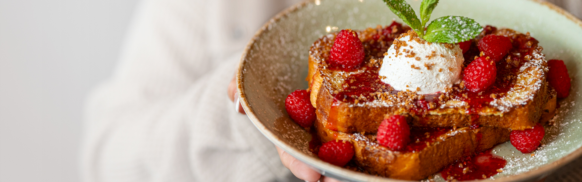 A close-up of an individual holding a plate of French Toast with raspberries, topped with ricotta and a mint sprig.